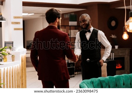 Diverse men shaking hands at hotel, african american concierge welcoming guest and offering his services. Bellhop provides excellent amenities to enhance business trip experience.
