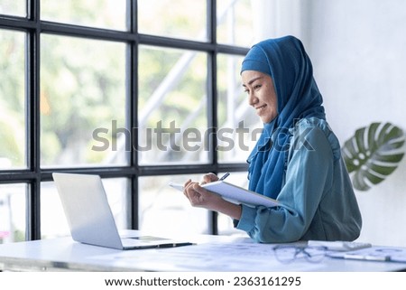 Business Asian muslim woman using calculator and writing make note with calculate doing math finance on an office desk. Woman working at office with laptop and tax, accounting, documents on desk
