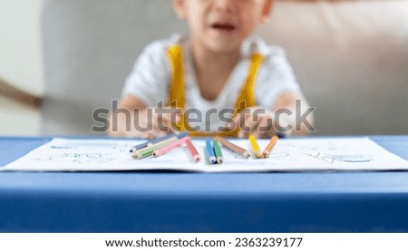Little sad boy is seating at table and crying with colorful pencils, indoors. Elementary school and education. Selective focus on subject.