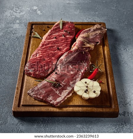 Square format, side perspective showcasing the diaphragm steak's detailed marbling. The beef, an alternative cut, lies on a wooden board with a gray setting. Royalty-Free Stock Photo #2363239613