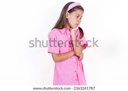Very bored Beautiful kid girl wearing pink dress over white background holding hand on cheek while support it with another crossed hand, looking tired and sick,