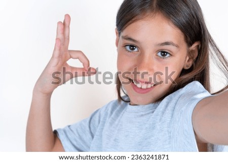 Portrait of pretty cheerful Beautiful kid girl wearing grey T-shirt over white background make selfie show okey symbol