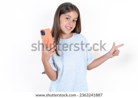 Astonished Beautiful kid girl wearing grey T-shirt over white background holding her telephone and pointing with finger aside at empty copy space