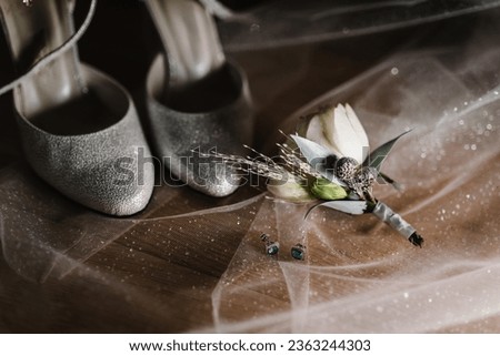 Wedding accessory bride. Stylish silver shoes, earrings, flowers, boutonniere, buttonhole on the table lying on background veil. Silhouette and shadow, sunshine. Closeup.