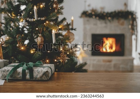 Stylish christmas gift close up against decorated tree and burning fireplace. Merry Christmas! Beautiful modern wrapped present with green ribbon on wooden table. Christmas background, copy space