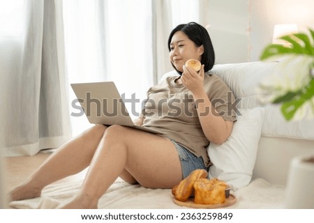 A happy Asian plus-size woman in comfy clothes is using her laptop computer and enjoying her yummy doughnuts in her bedroom. work from home, freelance, leisure, unhealthy lifestyle