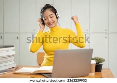 A cheerful and happy young Asian female college student enjoys listening to music on her wireless headphones while doing homework in a library.