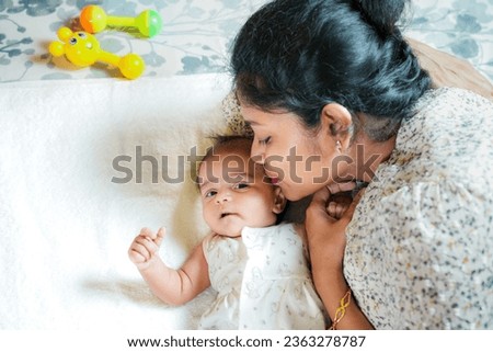 An Indian mother kissing her newborn baby girl on her forehead with love and affection while the cute adorable baby is looking to the camera. Few toys are also lying on the bed.