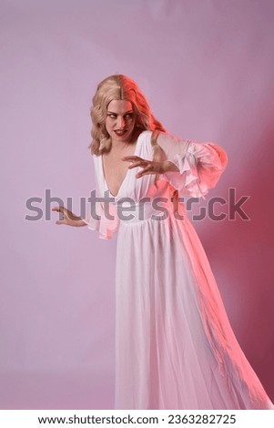 Close up portrait of beautiful blonde model wearing elegant  white halloween gown. arms reaching out like hungry vampire.  isolated on studio background with  red cinematic moody lighting.