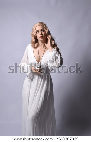 Close up portrait of beautiful blonde model wearing elegant  white halloween gown, a historical fantasy character.  
Holding crucifix cross necklace, isolated on studio background.