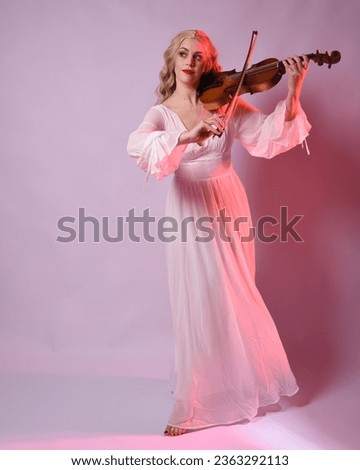 Full length portrait of beautiful blonde model wearing elegant white halloween gown, historical fantasy character.  facing backwards holding a violin musical instrument, isolated on studio background.