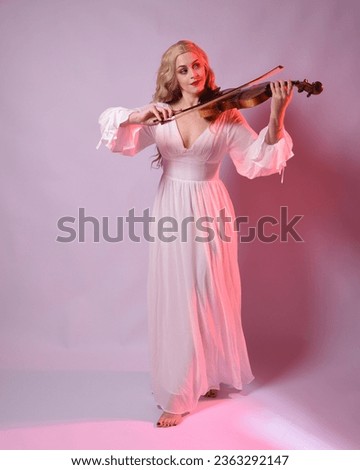 Full length portrait of beautiful blonde model wearing elegant white halloween gown, historical fantasy character.  facing backwards holding a violin musical instrument, isolated on studio background.