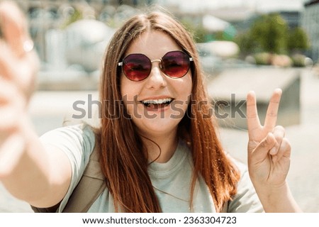 Lovely 30s girl in sunglasses posing with peace sign. Laughing young woman taking selfie on city background.