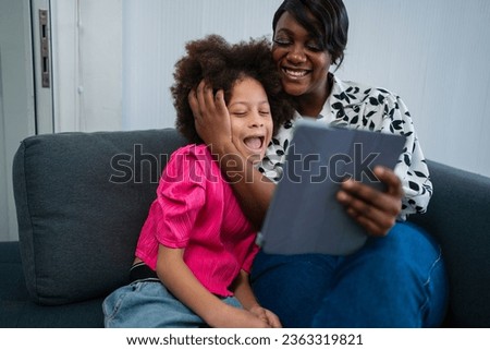 Young african american woman with cheerful and excited little girl using digital tablet on couch at home. Young black mother and smiling daughter playing on digital tablet at home. 