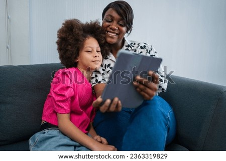 Young african american woman with cheerful and excited little girl using digital tablet on couch at home. Young black mother and smiling daughter playing on digital tablet at home. 