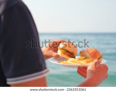 Man holds hamburger and french fries plate behind blurred sea view 