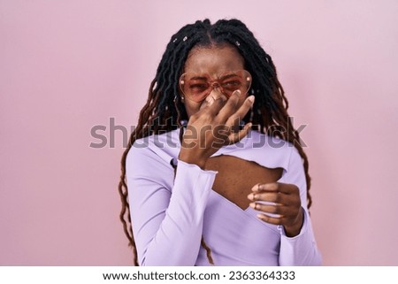 African woman with braided hair standing over pink background smelling something stinky and disgusting, intolerable smell, holding breath with fingers on nose. bad smell 