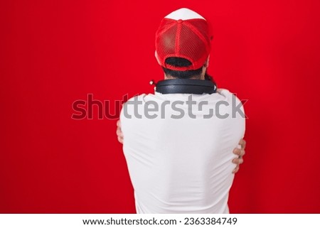 Hispanic man with beard wearing gamer hat and headphones hugging oneself happy and positive from backwards. self love and self care 
