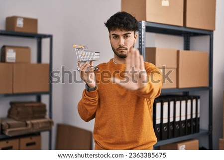 Hispanic man with beard standing by manikin at small business holding cart with open hand doing stop sign with serious and confident expression, defense gesture 