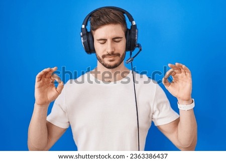 Hispanic man with beard listening to music wearing headphones relaxed and smiling with eyes closed doing meditation gesture with fingers. yoga concept.  Royalty-Free Stock Photo #2363386437