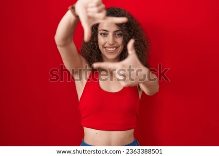 Hispanic woman with curly hair standing over red background smiling making frame with hands and fingers with happy face. creativity and photography concept. 