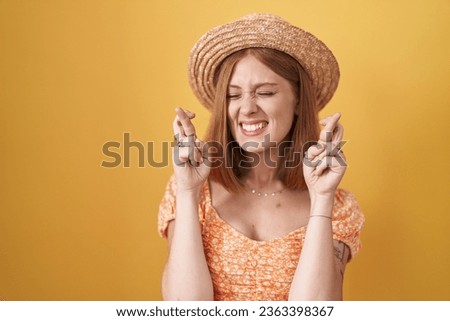 Young redhead woman standing over yellow background wearing summer hat gesturing finger crossed smiling with hope and eyes closed. luck and superstitious concept. 
