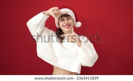 Young caucasian woman smiling doing frame shape with hands wearing christmas hat over isolated red background