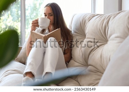 Young woman with cup of tea reading book on couch in living room Royalty-Free Stock Photo #2363430149