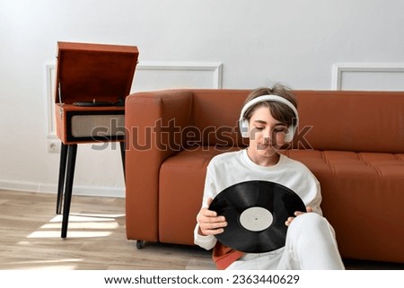Teenager boy with headphones holding in hands vinyl record and looking on it. Brown wooden turntable and sofa on background, home interior, lifestyle.