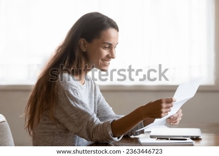 Smiling young woman reading pleasant news in letter, holding paper sheet, checking post mail, sitting at table at home, happy girl received refund, great shopping offer or exam results Royalty-Free Stock Photo #2363446723