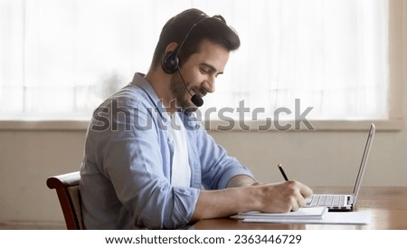 Smiling young man wearing headset with microphone writing notes, sitting at work desk with laptop, student listening to lecture, watching webinar, online course, e-learning, customer support service