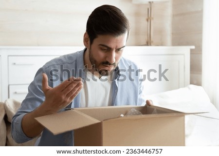Angry irritated man unboxing parcel, looking into open cardboard box, sitting on couch at home, dissatisfied annoyed customer received wrong or broken online store order, bad delivery service concept Royalty-Free Stock Photo #2363446757