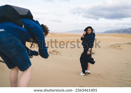 Full body of young mixed race female traveler with smart watch standing with hand touching hair in desert sand while looking at camera and posing to unrecognizable male with backpack shooting pictures