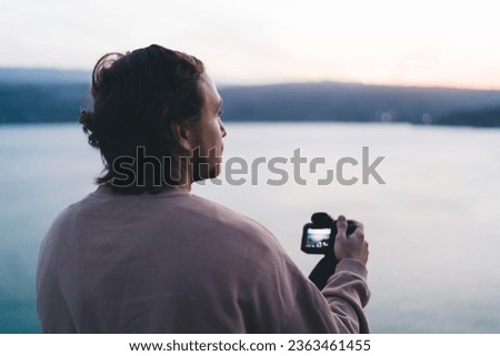 Back view of relaxing male photographer with professional camera in hands with pictures on screen and looking away while taking snaps of panoramic view of blurred sea mountains during sunset