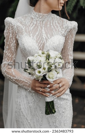 The bride in a white wedding dress embroidered with stones and beads holds a wedding bouquet of roses and greenery. Stylish wedding bouquet.