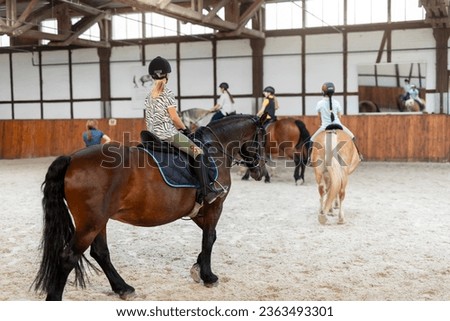 Horse riding school. Little children girls at group training equestrian lessons in indoor ranch horse riding hall. Cute little beginner blond girl kid in helmet sitting on brown horse horseback Royalty-Free Stock Photo #2363493301