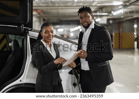 Attractive african woman signing digital contract online via tablet while standing near EV in underground parking. Business partners enhancing document accuracy while powering modern electric auto.