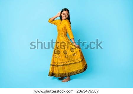 Full Length Of Smiling Young Indian Lady Wearing Beautiful Yellow Traditional Dress With Golden Embroidery And Strass, Dancing While Posing Over Blue Background, Studio Shot Royalty-Free Stock Photo #2363496387