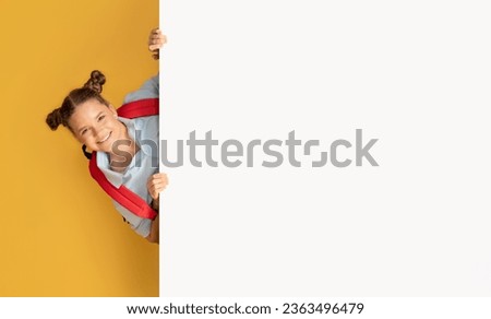 Happy european teenager schoolgirl looking from behind large banner with free space at school, isolated on an orange studio background. Sale, recommendation for study, knowledge, ad and offer