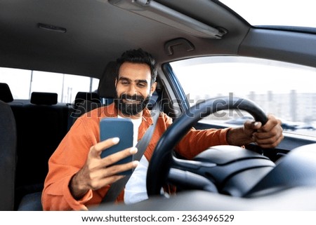 Cheerful arab driver man texting on smartphone while driving sitting in modern car inside. Man using mobile phone with gps navigator application navigating his new automobile, selective focus Royalty-Free Stock Photo #2363496529