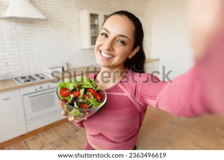 Happy fitness woman making selfie holding fresh vegetable salad bowl, smiling at camera, enjoying her weight loss routine and eating healthy food on a diet, posing at home kitchen. Selective focus