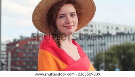 Portrait of a beautiful young woman in a hat with long red hair covering herself in lgbt flag while looking to camera. Alone. One. Keeping fist up, covering LGBT flag.