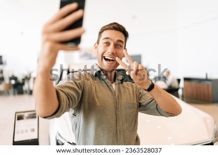 Portrait of cheerful male blogger streaming online from car dealership reviewing new auto holding smartphone. Front view of friendly young man vlogging selecting automobile at showroom.