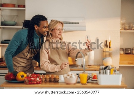 A cheerful interracial couple is cooking a healthy meal together at home while standing in a kitchen and taking self-portraits for social media. A couple is posting pictures on social networks.