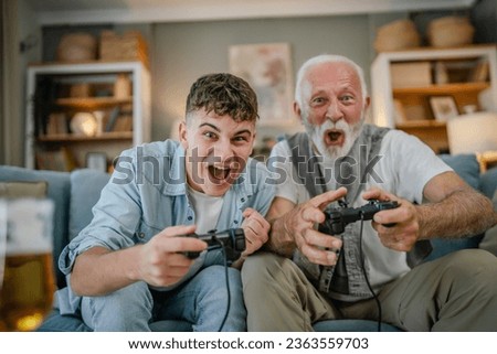 Caucasian teenager and senior man grandfather and grandson sit on sofa bed at home play console video game hold joystick controller have fun family bonding males men