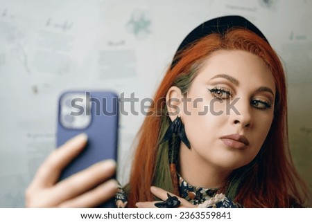 Female blogger with bright makeup. Red-haired girl takes selfie on front camera of smartphone. Woman with Gothic-style jewelry with proud expression on her face. Witch aesthetic.