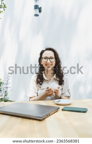 Young European woman sitting at outdoor cafe table with laptop and cup of coffee, smiling woman in glasses enjoying telecommuting in cafe or studying online Royalty-Free Stock Photo #2363568381