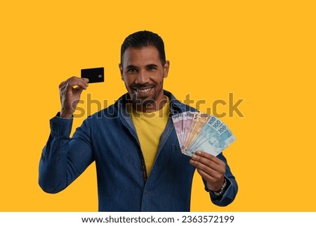 man holds a black credit card and Brazilian money, wearing blue and yellow clothes on a yellow background