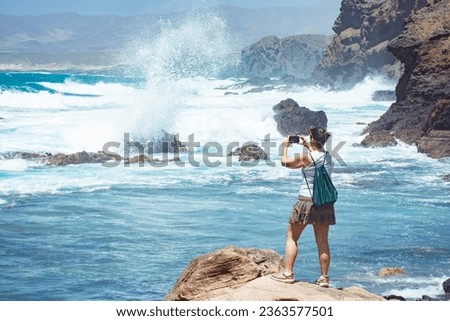 White mature woman standing on a rock taking photos with her smartphone of the sea landscape with waves from the strong wind in Fuerteventura with mountains in the background and clear blue sky.