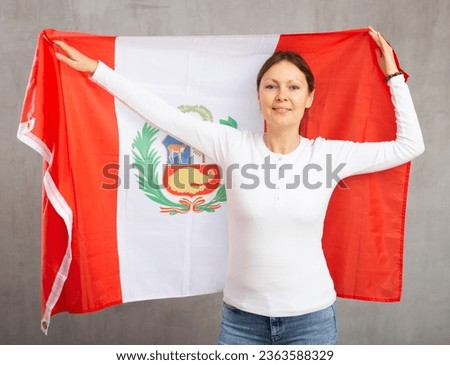 Happy young woman with large flag of Peru posing gladly against light unicoloured background